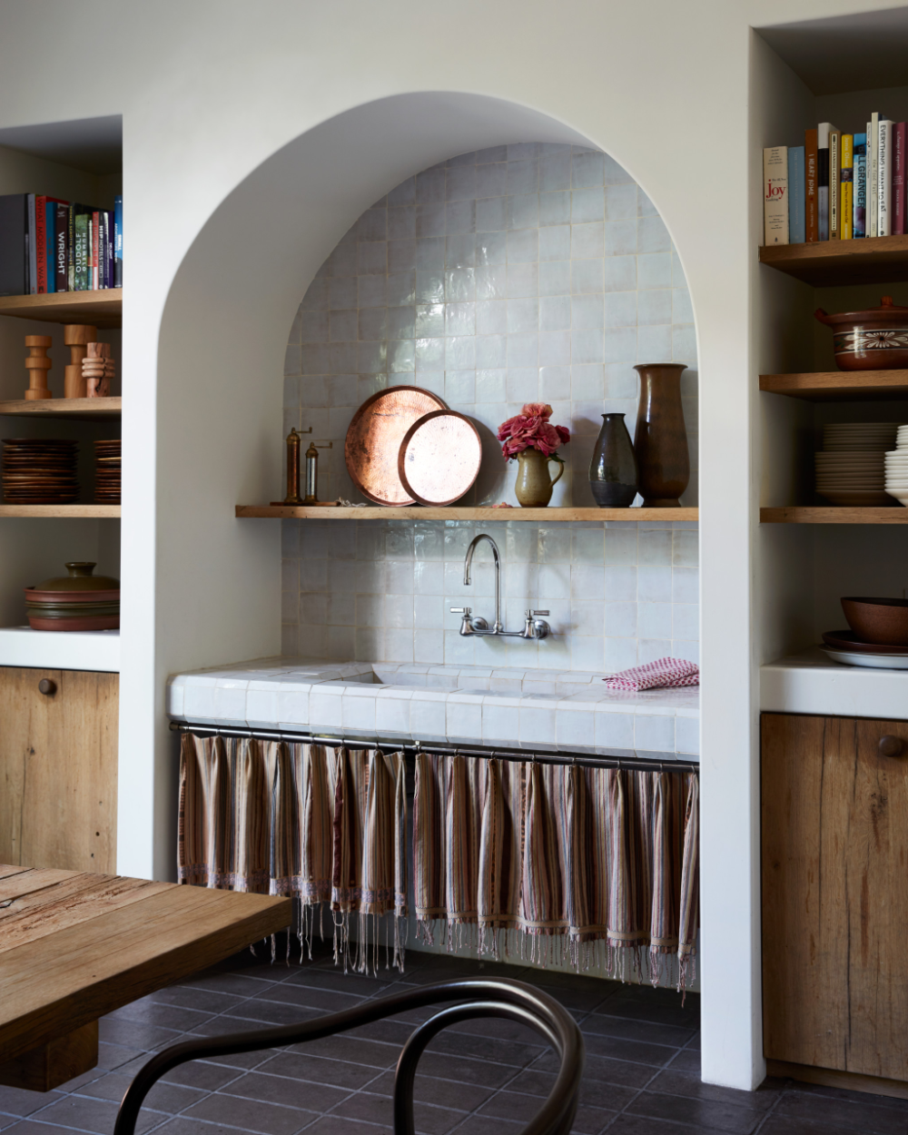 A modern rustic kitchen nook featuring a tiled countertop with a striped silk fabric skirt hanging underneath, concealing storage. The wooden shelf above holds decorative pottery and copper trays. The surrounding cabinets and shelves are made of natural wood, creating a warm and inviting atmosphere. Interior designer in Sacramento knows how to create a kitchen that works